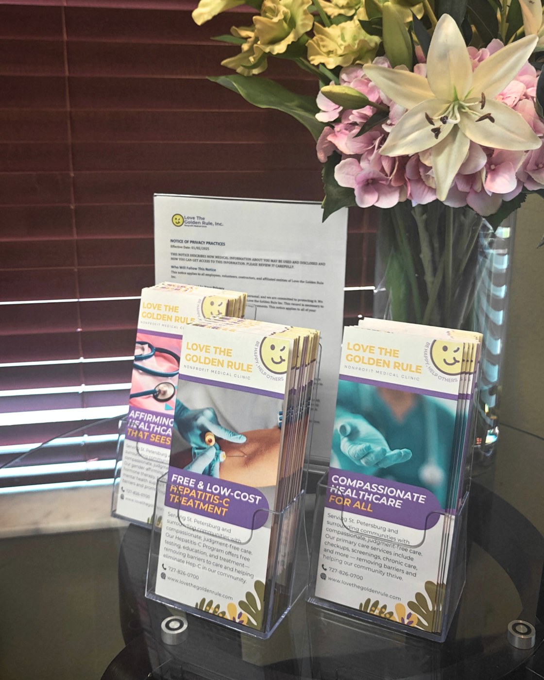 Clinic lobby table with Love The Golden Rule brochures for affirming healthcare, free Hepatitis-C treatment, and compassionate primary care, beside a vase of lilies