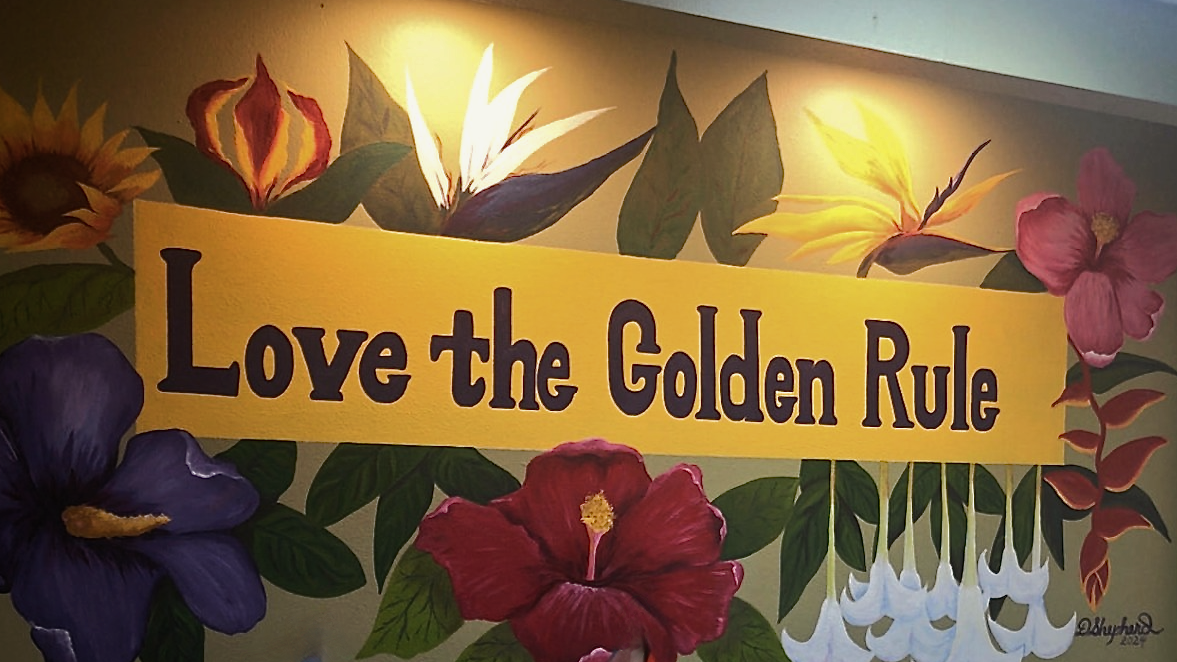 Hand-painted lobby mural reading “Love the Golden Rule,” surrounded by tropical flowers, with pride and transgender flags on the front desk